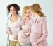 Three happy and proud, pregnant young adult women standing in a row against white background, looking at each other and smiling.
